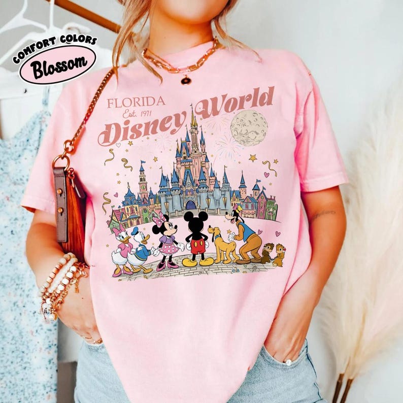 Puede incluir: Camiseta rosa claro con un dise&ntilde;o de Disney World en Florida. El dise&ntilde;o incluye el castillo de Disney World, fuegos artificiales y personajes de Disney. La camiseta tambi&eacute;n presenta el texto "FLORIDA Est. 1971 Disney World" en un estilo vintage.