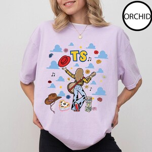 May include: A light purple t-shirt featuring a graphic print of a person with a guitar, a red hat, a brown cowboy hat, blue clouds, and the text TS. The design includes small flowers, musical notes, and stacked blocks with letters.