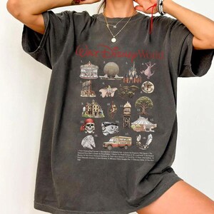 Puede incluir: Camiseta gris oscuro con un gr&aacute;fico detallado de varias atracciones de Walt Disney World, incluyendo el Castillo de Cenicienta y Spaceship Earth. Las palabras "Walt Disney World" est&aacute;n impresas en rojo en la parte superior.