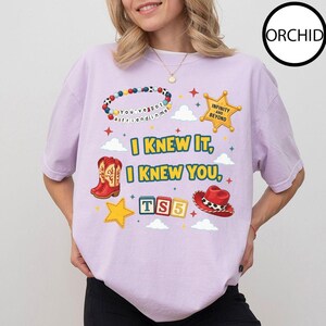 May include: A light purple t-shirt featuring a graphic print with a colorful beaded bracelet, a yellow sheriff star badge, red cowboy boots, a red cowboy hat, and alphabet blocks spelling TS5. The text reads I KNEW IT, I KNEW YOU, and INFINITY AND BEYOND.