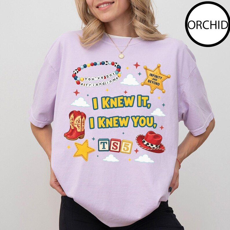 May include: A light purple t-shirt featuring a graphic print with a colorful beaded bracelet, a yellow sheriff star badge, red cowboy boots, a red cowboy hat, and alphabet blocks spelling TS5. The text reads I KNEW IT, I KNEW YOU, and INFINITY AND BEYOND.