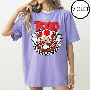 May include: A violet short-sleeve t-shirt featuring a graphic of the character Toad from Super Mario. The design includes the word TOAD in red letters above a circular black and white checkered border, red accents, and white lightning bolt shapes.