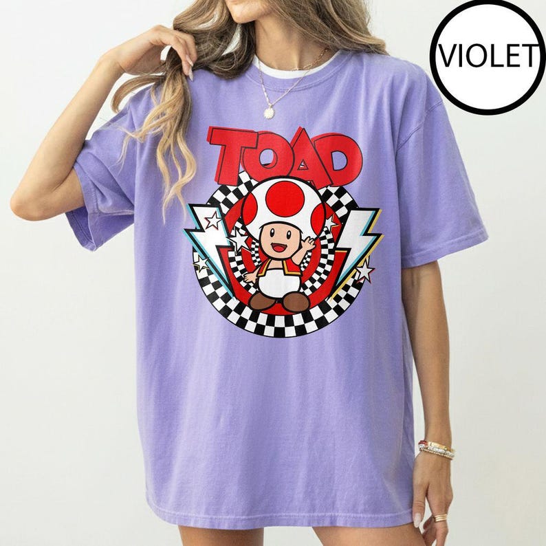May include: A violet short-sleeve t-shirt featuring a graphic of the character Toad from Super Mario. The design includes the word TOAD in red letters above a circular black and white checkered border, red accents, and white lightning bolt shapes.