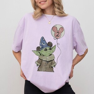 May include: A light purple t-shirt featuring a cartoon image of a green alien character wearing a wizard hat and holding a balloon with the number 2. The balloon is pink and white. The character is wearing a brown coat.