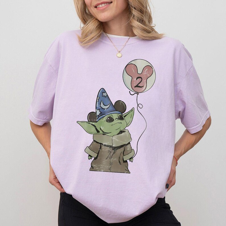 May include: A light purple t-shirt featuring a cartoon image of a green alien character wearing a wizard hat and holding a balloon with the number 2. The balloon is pink and white. The character is wearing a brown coat.