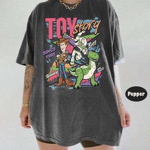 Puede incluir: Camiseta gris oscuro con un gr&aacute;fico colorido de Toy Story. El dise&ntilde;o presenta a Woody, Buzz Lightyear y Rex, junto con el texto "TOY Story" y "Since 1995".