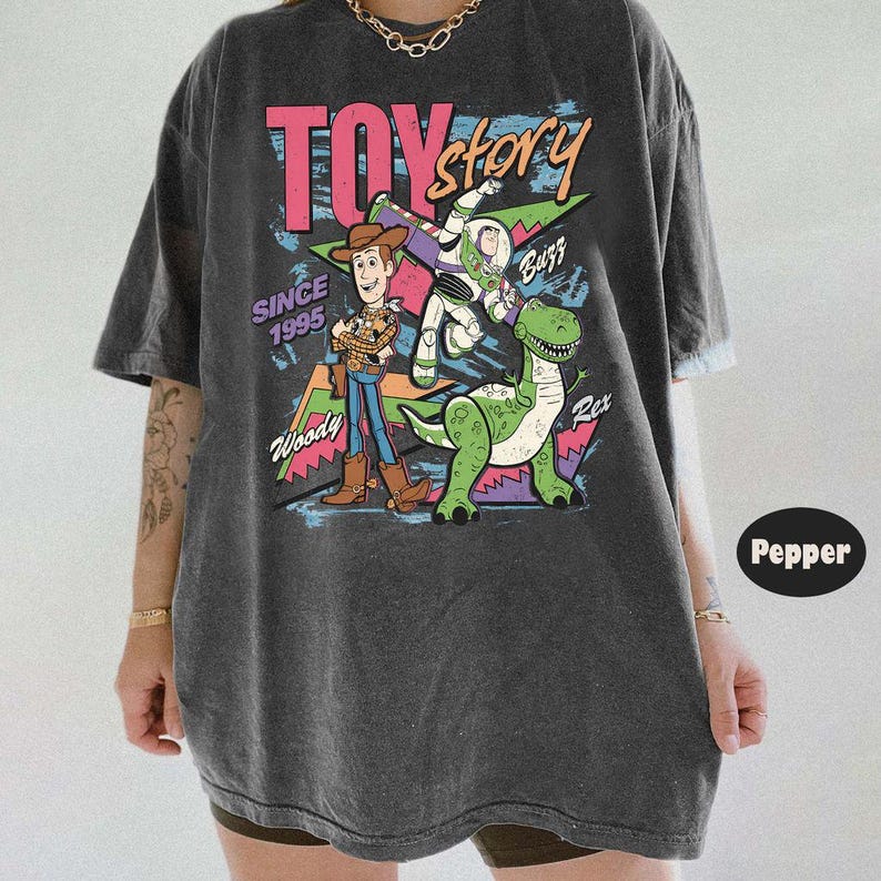 Puede incluir: Camiseta gris oscuro con un gr&aacute;fico colorido de Toy Story. El dise&ntilde;o presenta a Woody, Buzz Lightyear y Rex, junto con el texto "TOY Story" y "Since 1995".