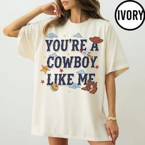 Puede incluir: Una camiseta de manga corta color marfil con el texto azul oscuro "YOU'RE A COWBOY LIKE ME" en el pecho. El gr&aacute;fico incluye ilustraciones de un sombrero de vaquero marr&oacute;n, un cohete, una estrella de sheriff amarilla, una pelota y estrellas rojas.