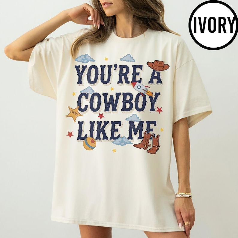 Puede incluir: Una camiseta de manga corta color marfil con el texto azul oscuro "YOU'RE A COWBOY LIKE ME" en el pecho. El gr&aacute;fico incluye ilustraciones de un sombrero de vaquero marr&oacute;n, un cohete, una estrella de sheriff amarilla, una pelota y estrellas rojas.
