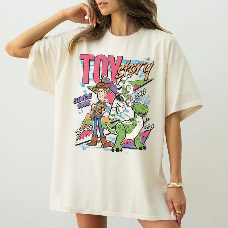 Puede incluir: Camiseta extragrande de color crema con un gr&aacute;fico colorido de los personajes de Toy Story. El dise&ntilde;o incluye a Woody, Buzz Lightyear y Rex, con las palabras "Toy Story" y "Since 1995". La camiseta tiene mangas cortas y un corte holgado.