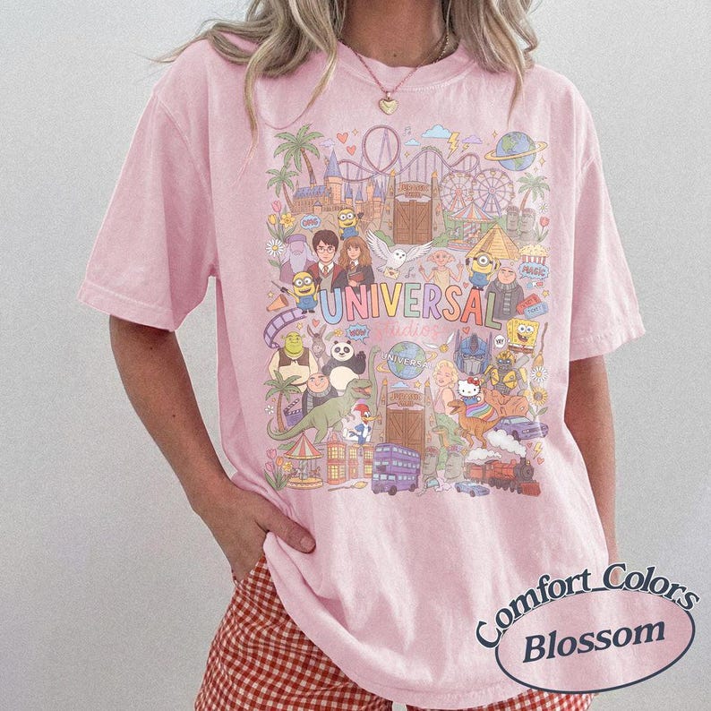 Może przedstawiać: Jasnor&oacute;żowy t-shirt z kr&oacute;tkim rękawem z kolorowym nadrukiem graficznym przedstawiającym postacie i punkty orientacyjne Universal Studios. Wz&oacute;r zawiera r&oacute;żne animowane postacie, kolejkę g&oacute;rską oraz napis UNIVERSAL dużymi, wielokolorowymi literami.
