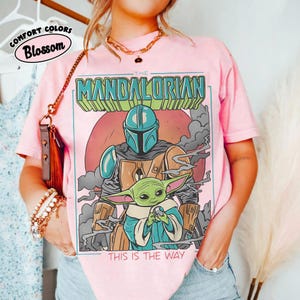 May include: Pink t-shirt featuring a graphic of The Mandalorian and Grogu, with the text "THIS IS THE WAY." The design includes a helmeted figure and a green alien character. The shirt also has the text "COMFORT COLORS Blossom" in a circle.