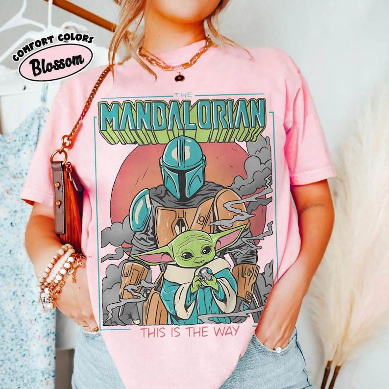 May include: Pink t-shirt featuring a graphic of The Mandalorian and Grogu, with the text "THIS IS THE WAY." The design includes a helmeted figure and a green alien character. The shirt also has the text "COMFORT COLORS Blossom" in a circle.