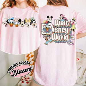Pu&ograve; includere: T-shirt rosa chiaro con personaggi Disney sul davanti e sul retro. Il davanti presenta una fila di personaggi, mentre il retro mostra la scritta "Walt Disney World" con un design arcobaleno e altri personaggi Disney.