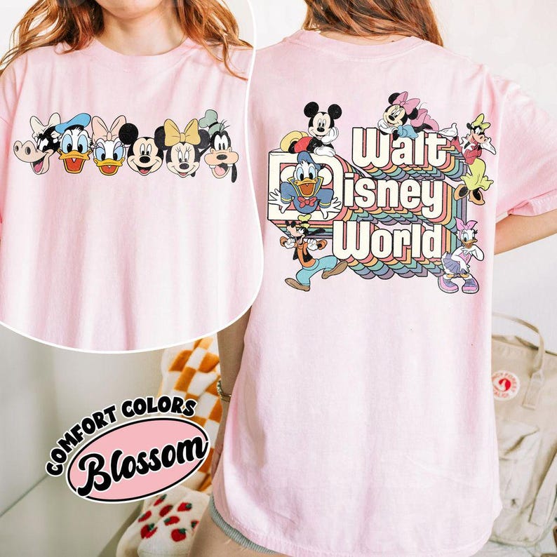 Pu&ograve; includere: T-shirt rosa chiaro con personaggi Disney sul davanti e sul retro. Il davanti presenta una fila di personaggi, mentre il retro mostra la scritta "Walt Disney World" con un design arcobaleno e altri personaggi Disney.