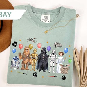 May include: A light green t-shirt featuring Star Wars characters holding balloons. The graphic includes R2-D2, Chewbacca, C-3PO, Darth Vader, a Stormtrooper, Princess Leia, and Grogu. The word "BAY" is in a white banner in the upper left.