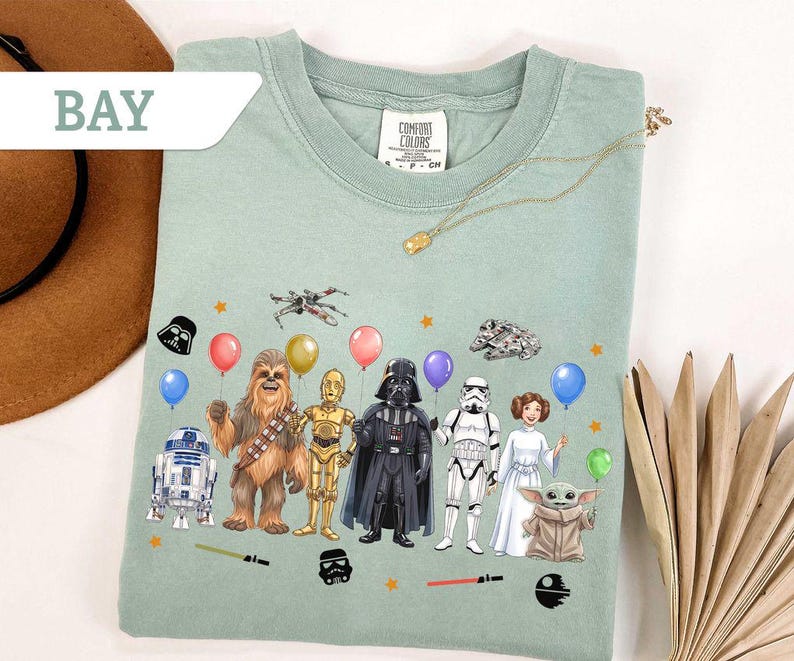 May include: A light green t-shirt featuring Star Wars characters holding balloons. The graphic includes R2-D2, Chewbacca, C-3PO, Darth Vader, a Stormtrooper, Princess Leia, and Grogu. The word "BAY" is in a white banner in the upper left.