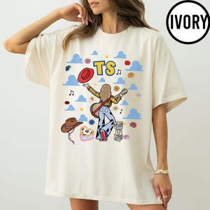 May include: An ivory t-shirt featuring a graphic of a person with long hair playing a guitar, wearing cow-print pants. The design includes blue clouds, red hats, flowers, musical notes, and blocks with the text TS. The shirt has a round neck.