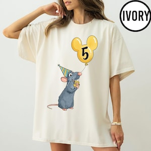 May include: Ivory-colored t-shirt featuring a cartoon rat wearing a party hat and holding a piece of cheese. A yellow balloon shaped like Mickey Mouse ears displays the number 5. The word "IVORY" is in a black circle.
