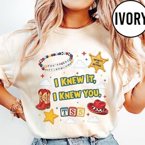 May include: An ivory t-shirt featuring a graphic print with a beaded bracelet reading "You've got a friend in me," a gold sheriff star badge, red cowboy boots, a red cowboy hat, and blocks spelling "TS5." Text reads "I KNEW IT, I KNEW YOU."