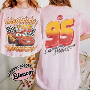 K&ouml;nnte beinhalten: Hellrosa T-Shirt mit Lightning McQueen aus dem Film Cars. Auf der Vorderseite ist McQueen mit der Nummer 95, Zielflaggen und Text abgebildet. Auf der R&uuml;ckseite steht gro&szlig; '95' und 'Lightning McQueen'.