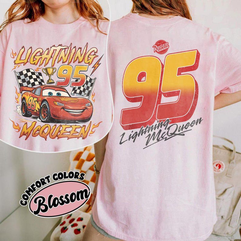 K&ouml;nnte beinhalten: Hellrosa T-Shirt mit Lightning McQueen aus dem Film Cars. Auf der Vorderseite ist McQueen mit der Nummer 95, Zielflaggen und Text abgebildet. Auf der R&uuml;ckseite steht gro&szlig; '95' und 'Lightning McQueen'.