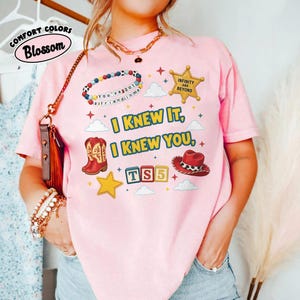 May include: A light pink short-sleeve t-shirt featuring a graphic print with the text "I KNEW IT, I KNEW YOU, TS5". The design includes a beaded bracelet, a gold sheriff star, red cowboy boots, a red cowboy hat, and small yellow stars.