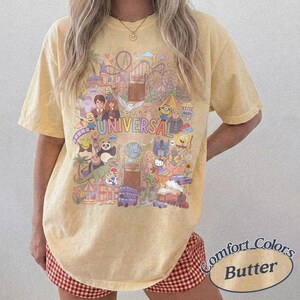 Może przedstawiać: Maślanie ż&oacute;łty t-shirt z kr&oacute;tkim rękawem z kolorowym nadrukiem graficznym przedstawiającym postacie i atrakcje Universal Studios. Wz&oacute;r zawiera r&oacute;żne animowane postacie, kolejkę g&oacute;rską oraz napis Universal Studios wielokolorową czcionką.