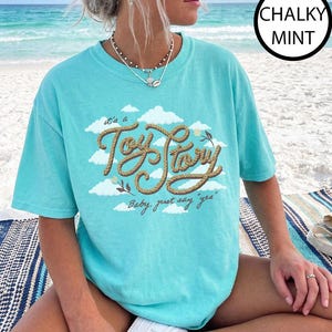 May include: A chalky mint short-sleeve t-shirt featuring a graphic of white clouds and the text "it's a Toy Story" written in a brown rope-style font, with the additional text "Baby, just say "yes"" printed below in a cursive brown script.