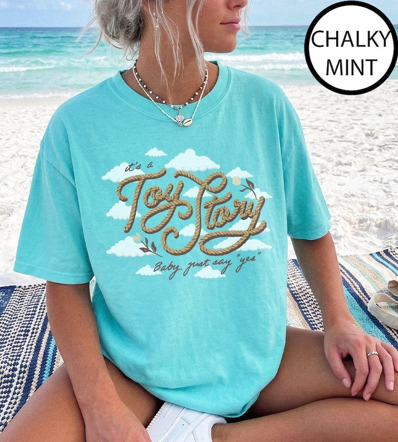 May include: A chalky mint short-sleeve t-shirt featuring a graphic of white clouds and the text "it's a Toy Story" written in a brown rope-style font, with the additional text "Baby, just say "yes"" printed below in a cursive brown script.