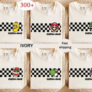May include: Ivory t-shirts with cartoon characters like Mario and Peach, each wearing sunglasses. A black and white chequered stripe and the text "CUSTOM NAME" are featured on each shirt. The image also includes the text "300+" and "Fast shipping".