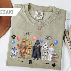 May include: Khaki-colored t-shirt featuring Star Wars characters with balloons and stars. The graphic includes R2-D2, Chewbacca, C-3PO, Darth Vader, a Stormtrooper, Princess Leia, and Grogu. The shirt has a Comfort Colors tag.