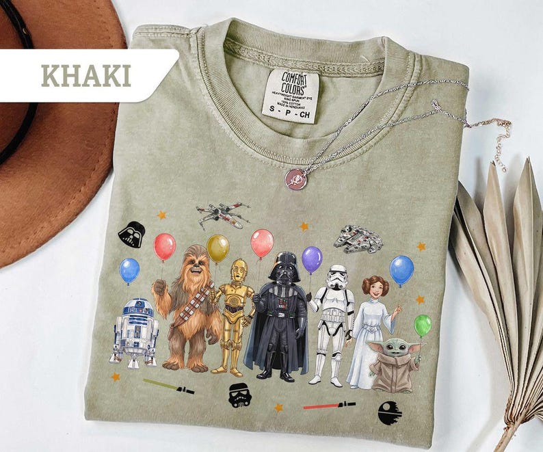 May include: Khaki-colored t-shirt featuring Star Wars characters with balloons and stars. The graphic includes R2-D2, Chewbacca, C-3PO, Darth Vader, a Stormtrooper, Princess Leia, and Grogu. The shirt has a Comfort Colors tag.