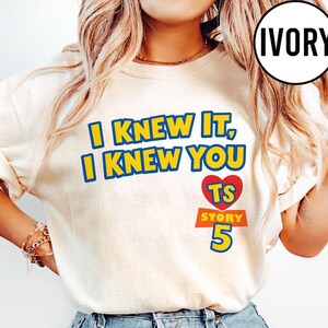 May include: An ivory t-shirt featuring the yellow text I KNEW IT, I KNEW YOU with blue outlines. Below the text is a red heart containing the yellow letters TS, followed by the orange text STORY and the large blue number 5 with a yellow outline.
