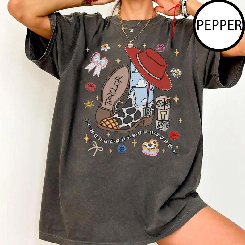 May include: A charcoal gray short-sleeve t-shirt featuring a graphic print of a cow-print cowboy boot with a red hat, blocks with the letters T, S, and 13, a bow, and the text TAYLOR. The design includes stars, flowers, and a beaded string.