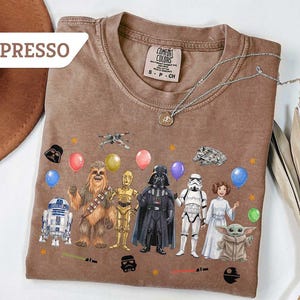May include: Espresso-colored t-shirt featuring Star Wars characters and balloons. The design includes R2-D2, Chewbacca, C-3PO, Darth Vader, a Stormtrooper, Princess Leia, and Grogu. The shirt has a Comfort Colors tag.
