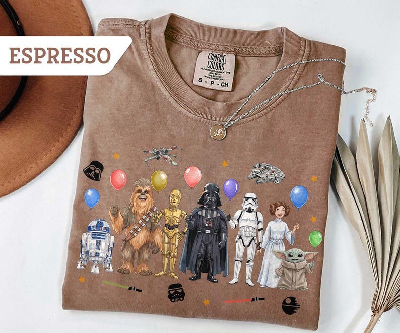 May include: Espresso-colored t-shirt featuring Star Wars characters and balloons. The design includes R2-D2, Chewbacca, C-3PO, Darth Vader, a Stormtrooper, Princess Leia, and Grogu. The shirt has a Comfort Colors tag.