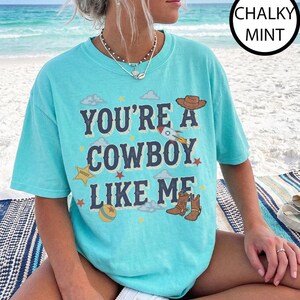 Puede incluir: Una camiseta de manga corta color menta tiza con el texto azul oscuro "YOU'RE A COWBOY LIKE ME" rodeado de ilustraciones gr&aacute;ficas de un sombrero de vaquero marr&oacute;n, un cohete, una estrella de sheriff amarilla, una pelota y botas vaqueras.