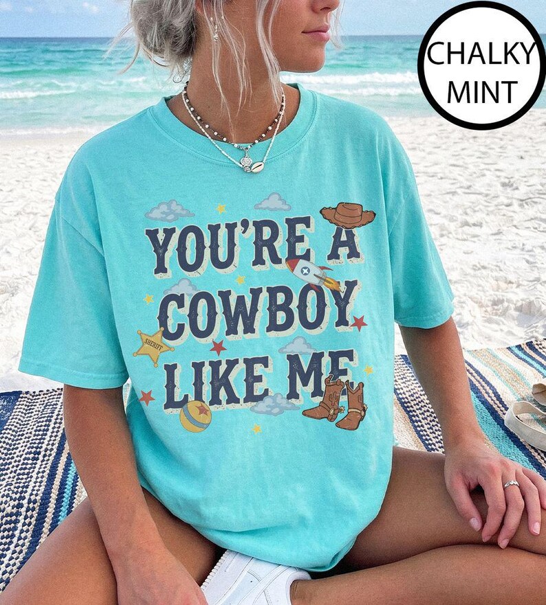 Puede incluir: Una camiseta de manga corta color menta tiza con el texto azul oscuro "YOU'RE A COWBOY LIKE ME" rodeado de ilustraciones gr&aacute;ficas de un sombrero de vaquero marr&oacute;n, un cohete, una estrella de sheriff amarilla, una pelota y botas vaqueras.