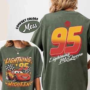 K&ouml;nnte beinhalten: Moosgr&uuml;nes T-Shirt mit Lightning McQueen aus dem Film Cars. Auf der Vorderseite ist McQueen mit der Nummer 95, einer Troph&auml;e und Zielflaggen abgebildet. Auf der R&uuml;ckseite steht eine gro&szlig;e 95 in Rot und Gelb mit "Lightning McQueen" darunter.