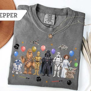 May include: Gray t-shirt featuring Star Wars characters and balloons. The design includes Darth Vader, Chewbacca, R2-D2, and other characters. The shirt has a crew neck and short sleeves. The word "PEPPER" is in a white banner.