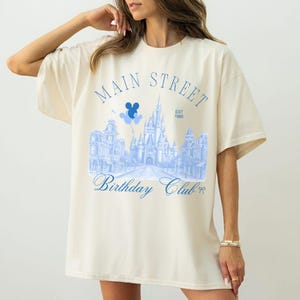 May include: Cream-colored oversized t-shirt featuring a light blue graphic of a castle, buildings, and the text "Main Street Birthday Club Est. 1986." The design includes Mickey Mouse balloons.
