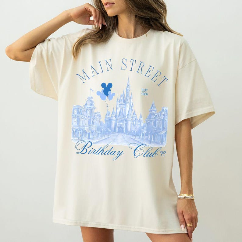 May include: Cream-colored oversized t-shirt featuring a light blue graphic of a castle, buildings, and the text "Main Street Birthday Club Est. 1986." The design includes Mickey Mouse balloons.