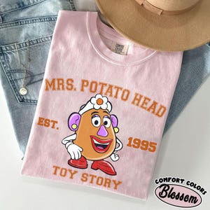 May include: A light pink t-shirt featuring a cartoon image of Mrs. Potato Head from Toy Story. The shirt has the text "MRS. POTATO HEAD" above the character, with "EST. 1995" and "TOY STORY" below. A tan hat and blue jeans are in the background.