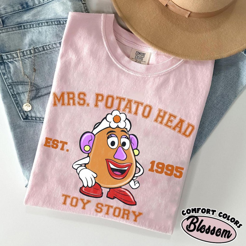 May include: A light pink t-shirt featuring a cartoon image of Mrs. Potato Head from Toy Story. The shirt has the text "MRS. POTATO HEAD" above the character, with "EST. 1995" and "TOY STORY" below. A tan hat and blue jeans are in the background.