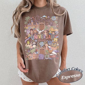 Może przedstawiać: Brązowy t-shirt z kr&oacute;tkim rękawem z kolorowym nadrukiem graficznym przedstawiającym postacie i atrakcje Universal Studios. Wz&oacute;r zawiera ilustracje Harry'ego Pottera, Shreka, Minionk&oacute;w oraz r&oacute;żnych element&oacute;w parku rozrywki z napisem Universal Studios.