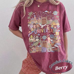 Może przedstawiać: Jagodowy t-shirt z kr&oacute;tkim rękawem z wielokolorowym nadrukiem graficznym przedstawiającym r&oacute;żne postacie animowane i zabytki Universal Studios. Wz&oacute;r zawiera napis Universal Studios oraz r&oacute;żnorodne postacie z bajek.