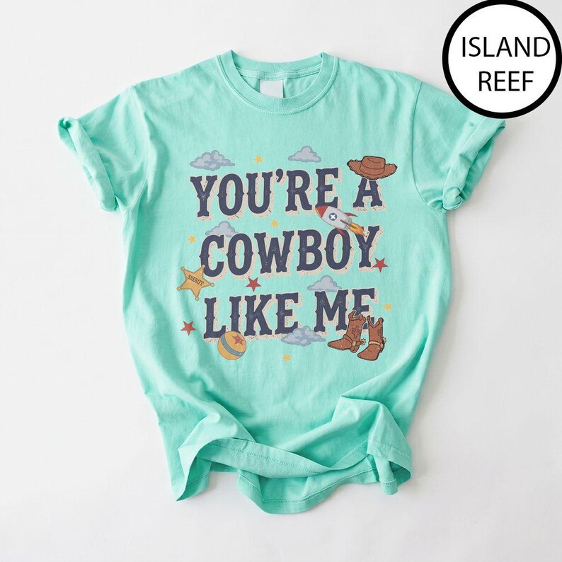 Puede incluir: Una camiseta de manga corta color verde menta con el texto "YOU'RE A COWBOY LIKE ME" en letras azul oscuro. El dise&ntilde;o incluye ilustraciones de un sombrero de vaquero marr&oacute;n, un cohete, una estrella de sheriff amarilla, una pelota y botas.