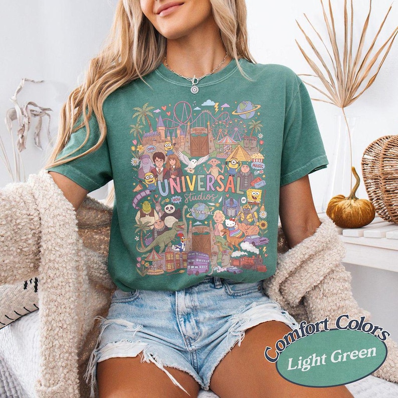 Może przedstawiać: Jasnozielony t-shirt z kolorowym nadrukiem graficznym przedstawiającym postacie i punkty orientacyjne Universal Studios, w tym Harry'ego Pottera, Shreka i Minionki, z napisem Universal Studios umieszczonym na środku wzoru.