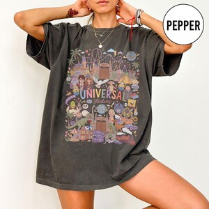 Może przedstawiać: Ciemnoszary t-shirt o kroju oversize z kolorowym nadrukiem graficznym przedstawiającym atrakcje i postacie z Universal Studios, w tym Harry'ego Pottera, Shreka i Minionki, z napisem Universal Studios na środku wzoru.
